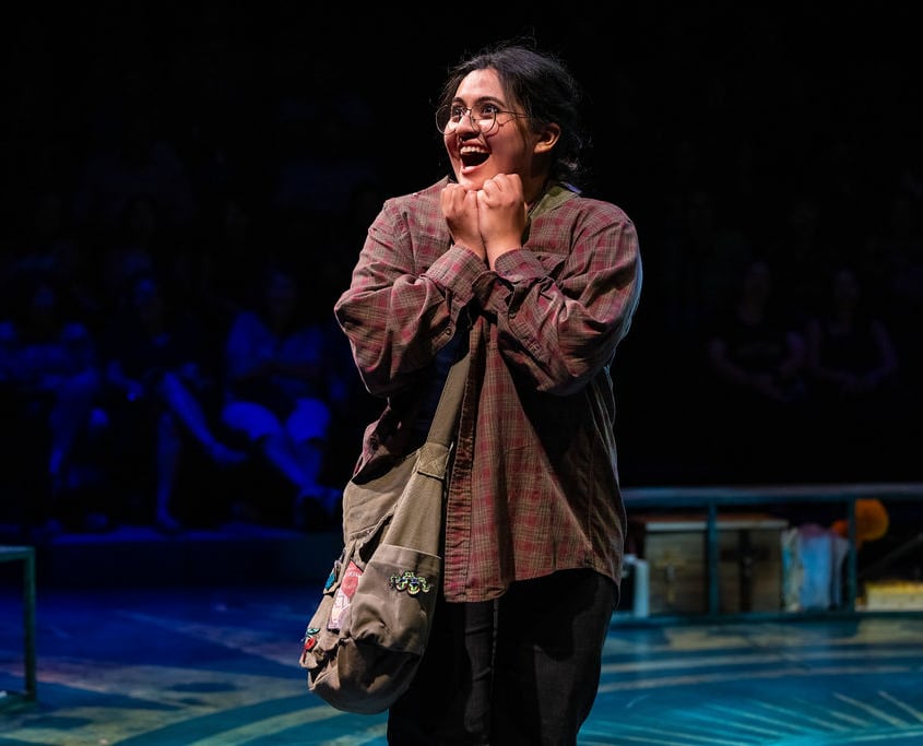 A teenage girl in a red and tan flannel and wearing round glasses grins excitedly