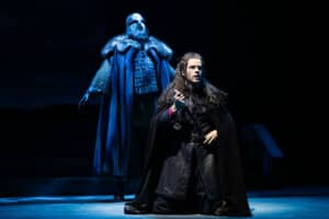 A young man wearing a black tunic and black cloak kneels and holding a dagger. A tall man is bathed in blue light, wearing armor, a leather breastplate, chain mail, and helmet, stands behind him.