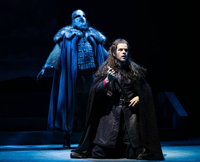 A young man wearing a black tunic and black cloak kneels and holding a dagger. A tall man is bathed in blue light, wearing armor, a leather breastplate, chain mail, and helmet, stands behind him.