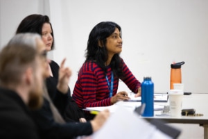 Playwright Vauhini Vara sits at a table with her peers. Papers and drinks are scattered across the table, the team is hard at work.