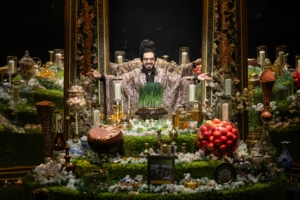 Michael Shayan playing his mother Roya is dressed in an intricate gold robe and large glasses, gestures grandly.