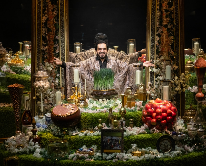 Michael Shayan playing his mother Roya is dressed in an intricate gold robe and large glasses, gestures grandly.