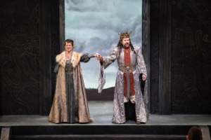 A man and woman wearing golden crowns clasp hands as they look out over the stage. The woman is wearing a green dress and a gold fur lined cloak. The man is wearing an orange tunic and a silver cloak with a large belt.