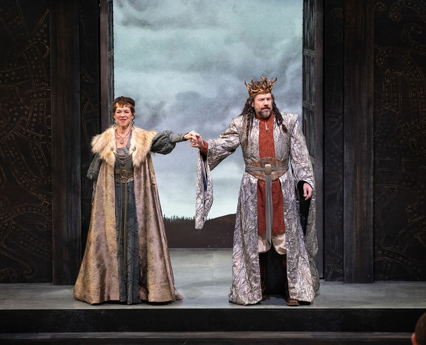 A man and woman wearing golden crowns clasp hands as they look out over the stage. The woman is wearing a green dress and a gold fur lined cloak. The man is wearing an orange tunic and a silver cloak with a large belt.