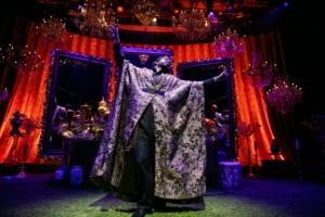 Michael Shayan playing his mother Roya is dressed in an intricate gold robe and large glasses, gestures grandly.