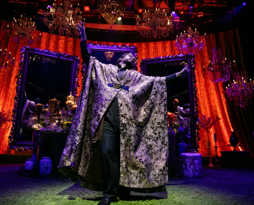 Michael Shayan playing his mother Roya is dressed in an intricate gold robe and large glasses, gestures grandly.