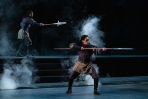 Two men dressed in tunics and leather armor hold out large spears, as they emerge from the fog.