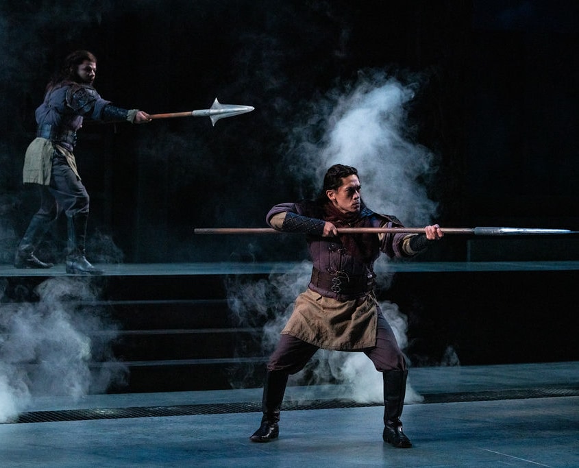 Two men dressed in tunics and leather armor hold out large spears, as they emerge from the fog.