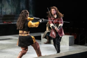 Two men with long braided hair wearing medieval tunics engage in an intense sword fight.