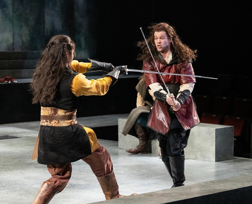 Two men with long braided hair wearing medieval tunics engage in an intense sword fight.