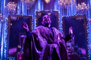 Michael Shayan playing his mother Roya is dressed in an intricate gold robe and large glasses, gestures grandly.