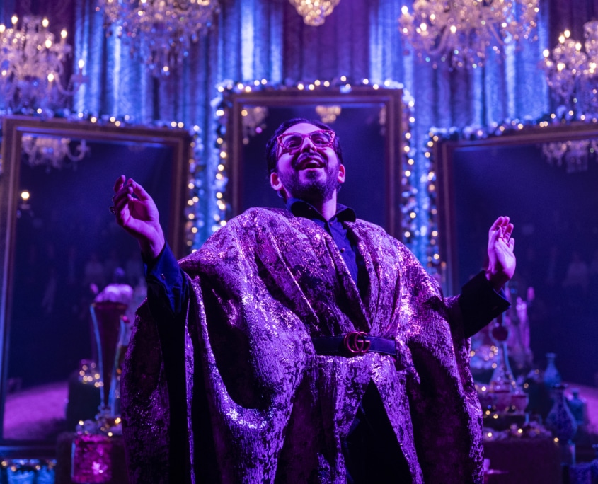 Michael Shayan playing his mother Roya is dressed in an intricate gold robe and large glasses, gestures grandly.