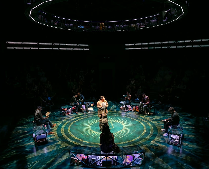 A teenage girl in a brown plaid blazer stands next to a suitcase holding a manuscript. She looks around the stage at the people sitting on benches around her, they are all reading I Am Not Your Perfect Mexican Daughter.