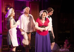 A group of performers sing onstage. They are wearing old-timey costumes and hats thrown on over their regular clothes