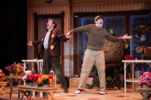 An older man in a brown cardigan and a young man wearing glasses and a baseball cap dance around a flower shop