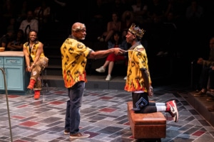 An older Black man knights a younger Black man with a large, slotted spoon, another Black man watches the scene, smiling. The young man is wearing a gold crown, all the men are wearing yellow and orange Hawiian shirts with flaming hearts on them.
