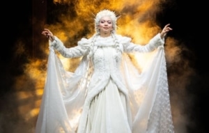 A woman, The Ghost of Christmas Past, regally spreads out her arms. She is wearing a long, intricate, white dress lined with white fur, a long white cape is connected to her wrists and spreads out behind her.