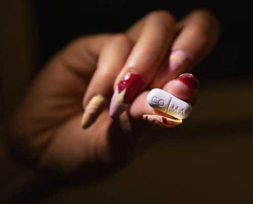 A woman’s hand with decorative acrylic nails holds a white pill towards the camera so that the word COMA printed on the pill is visible.
