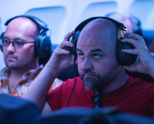 Two bald men sit in airplane seats, putting on large headphones. They look apprehensive.