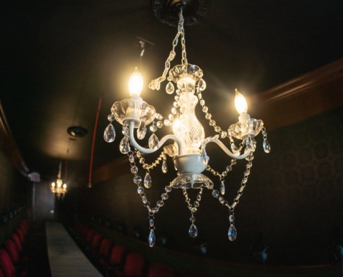 A lit crystal chandelier hangs above a long table. The table is lined with red velvet chairs and the walls of the room are covered in patterned green wallpaper