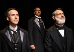 Two men stand in the foreground, another slightly behind. They're looking off into the distance. All are wearing old fashioned three piece suits.