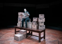 An older gentleman reads a newspaper. He's seated on a stack of wooden boxes, stacked on top of a wooden table.