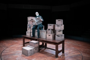An older gentleman reads a newspaper. He's seated on a stack of wooden boxes, stacked on top of a wooden table.