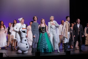 Olaf, Kristoff, Anna, Elsa, and Hans stand together, holding hands as they sing Let It Go onstage. A chorus in white stands behind them.