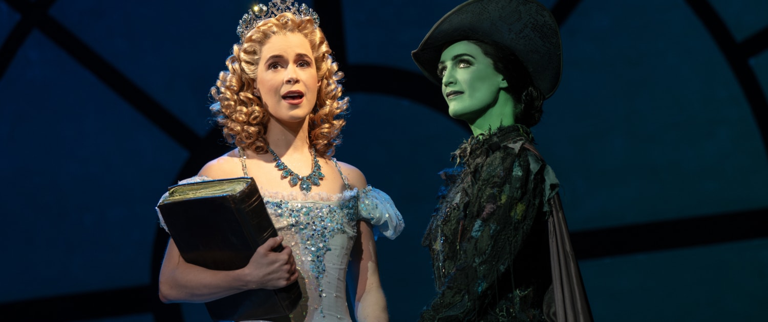 Glinda and Elphaba stand together, hands clasped. Glinda is holding an old tome.