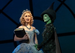 Glinda and Elphaba stand together, hands clasped. Glinda is holding an old tome.
