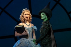 Glinda and Elphaba stand together, hands clasped. Glinda is holding an old tome.
