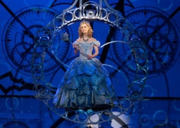 Glinda floats in her bubble down toward the stage. The bubble is comprised of little steel circles. She is wearing a blue, frilly ballgown.