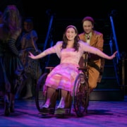 Boq pushes Nessarose in her wheelchair. She is smiling widely with her arms outstretched, glittering silver shoes on her feet.
