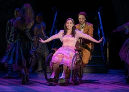 Boq pushes Nessarose in her wheelchair. She is smiling widely with her arms outstretched, glittering silver shoes on her feet.