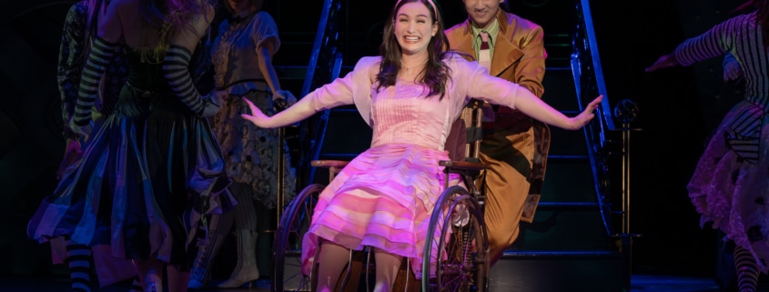 Boq pushes Nessarose in her wheelchair. She is smiling widely with her arms outstretched, glittering silver shoes on her feet.