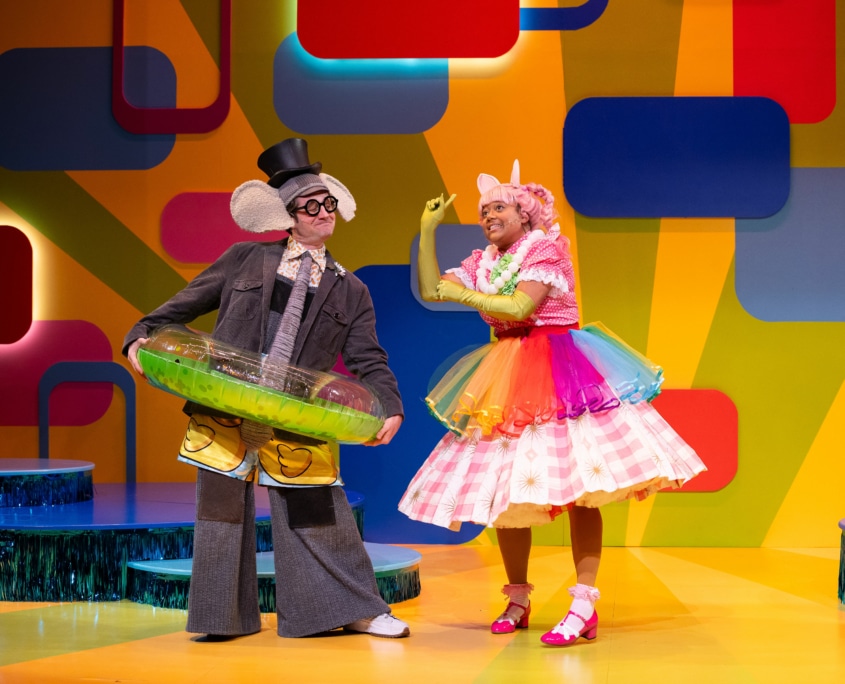 Gerald the Elephant is dressed in swim trunks, a top hat, and an inflatable swim ring on top of his normal clothes. Next to him Piggie is wearing a rainbow tutu, a flower necklace, and green satin gloves on top of her regular clothes.