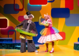 Gerald the elephant and Piggie stand onstage, wearing fun accessories like a pool inner tube, a rainbow tutu, and a top hat.