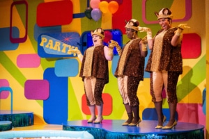 The three Squirrelles stand on a blue platform dancing. They are all wearing flowy sequined tops, pants, and hats.