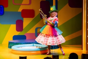 Piggie; dressed in a rainbow tutu, a flower necklace, green cowboy hat, and green satin gloves on top of her regular clothes holds up a trumpet