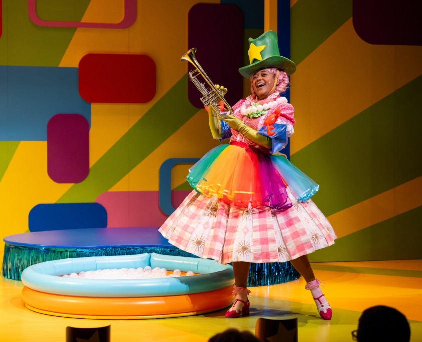 Piggie; dressed in a rainbow tutu, a flower necklace, green cowboy hat, and green satin gloves on top of her regular clothes holds up a trumpet
