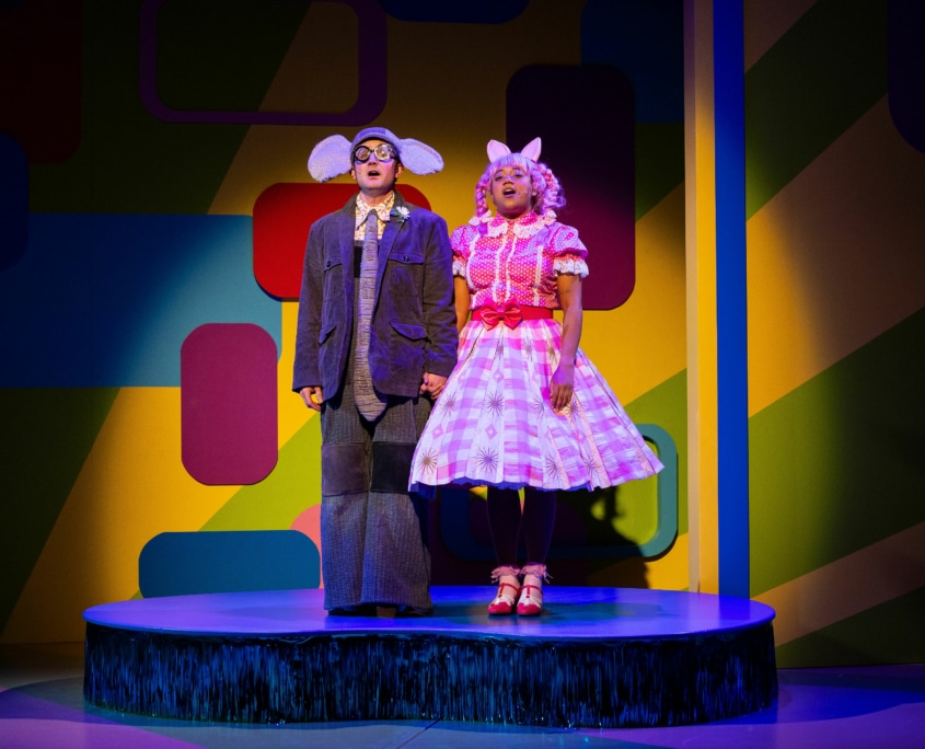 Gerald the Elephant and Piggie hold hands on top of a blue platform singing.