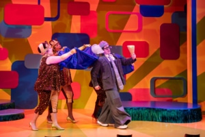 Gerald the Elephant marches across the stage wearing a blue cape and holding a giant ice cream cone. The three Squirrelles hold up his cape so that it blows behind him. Squirrelle