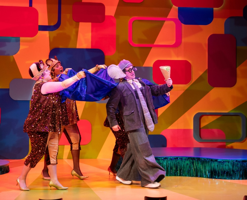 Gerald the Elephant marches across the stage wearing a blue cape and holding a giant ice cream cone. The three Squirrelles hold up his cape so that it blows behind him. Squirrelle